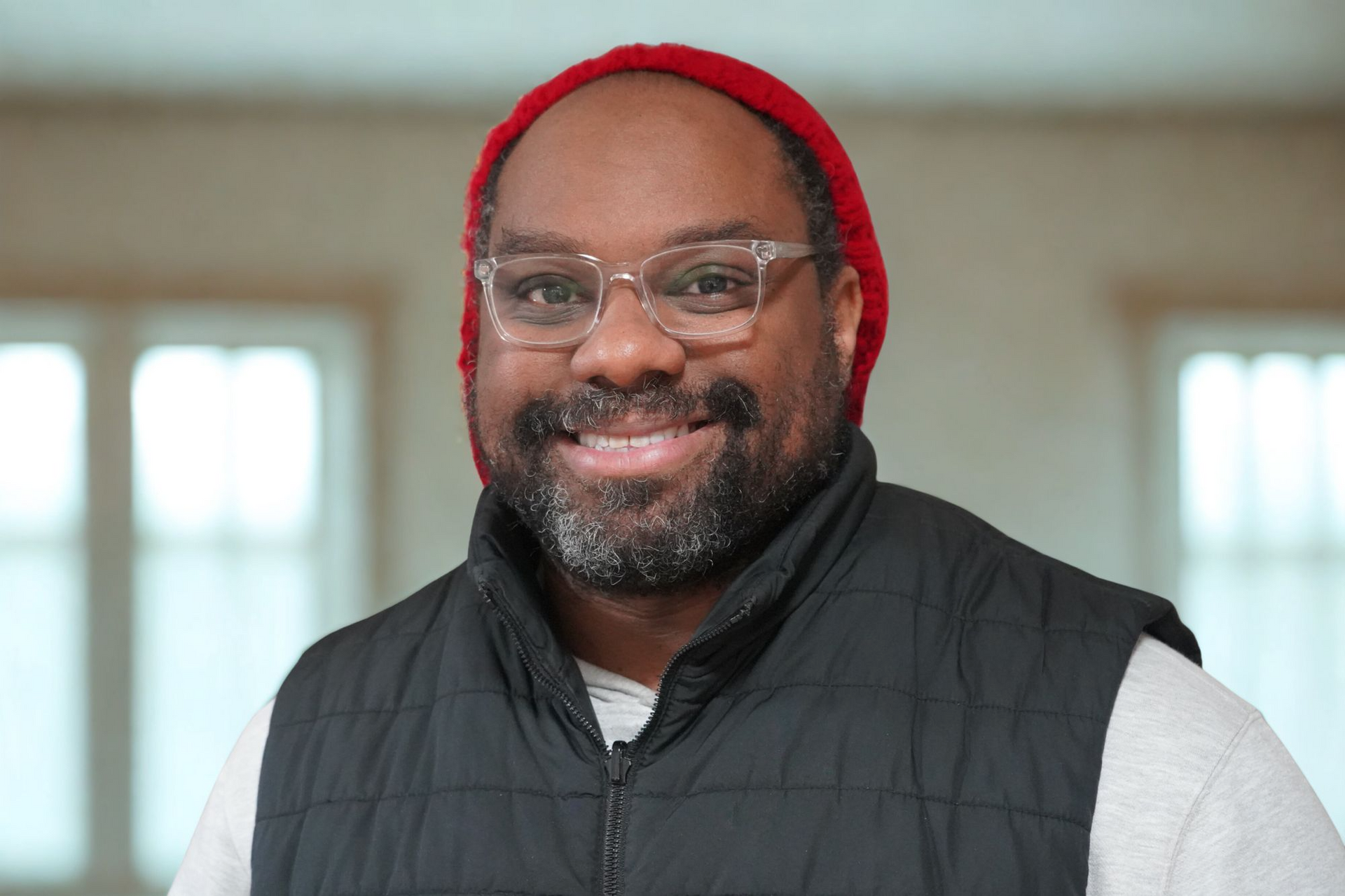 A smiling person wearing a red beanie, clear-framed glasses, and a black quilted vest over a light-colored shirt.