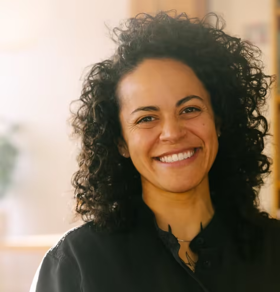 A person with curly dark hair and a bright, friendly smile, wearing a black shirt in a softly lit indoor setting.