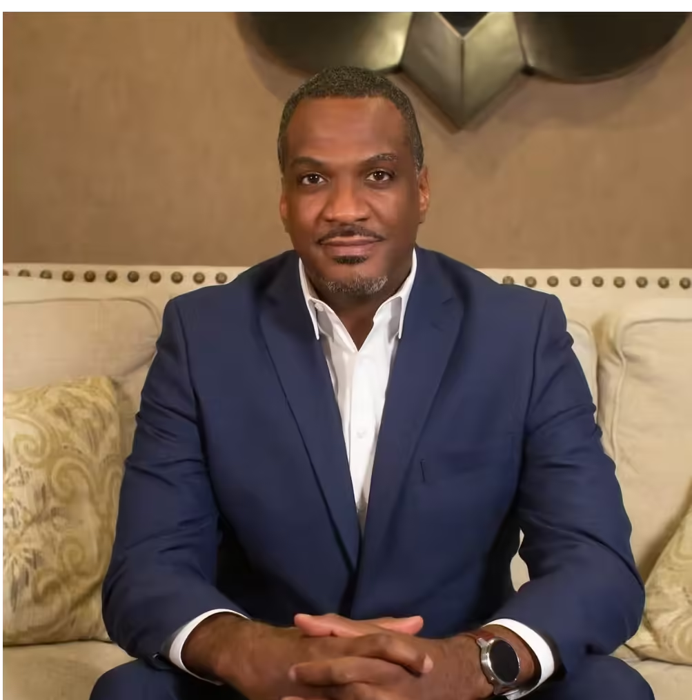 A person in a blue blazer and white dress shirt sits on a beige sofa, hands clasped, looking forward with a calm expression.