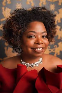 A person with curly, dark hair smiles in a red, off-the-shoulder ruffled top, wearing a crystal necklace against a patterned wall.
