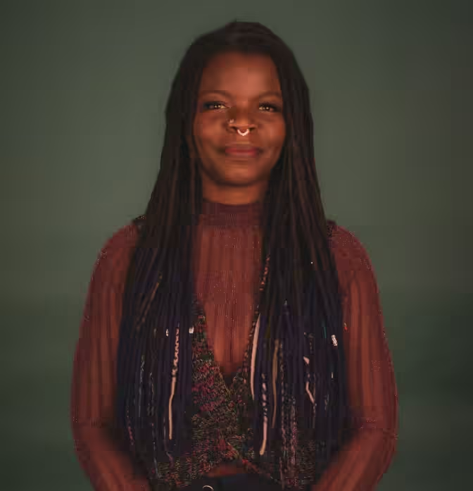A person with long, dark dreadlocks and a septum piercing, wearing a maroon textured top, centered against a muted background.