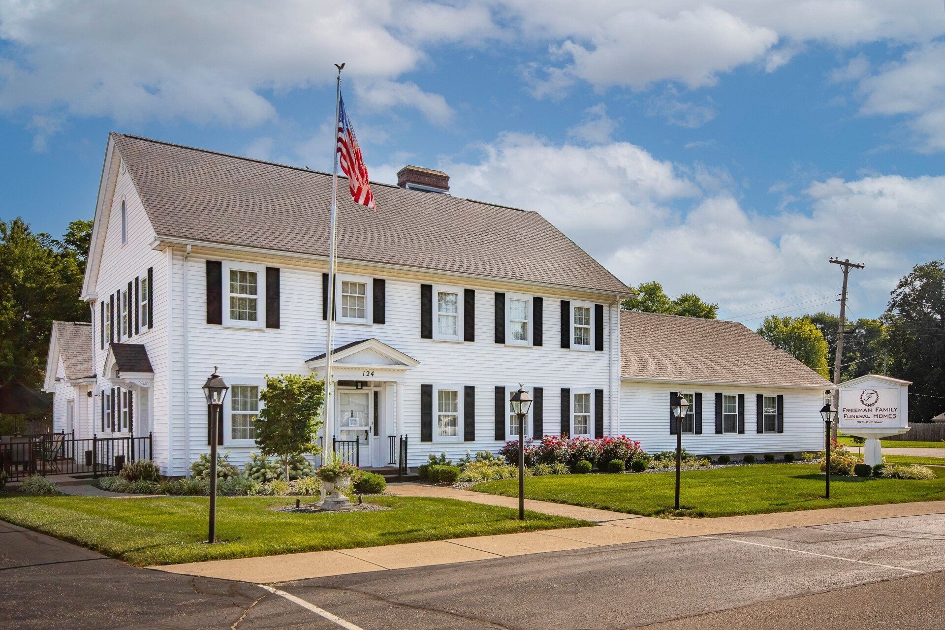 White two-story building with black shutters, an American flag, and a sign.