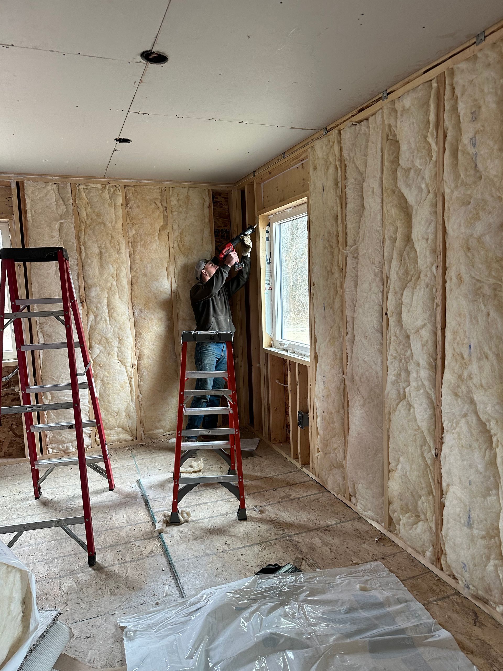 Interior room under renovation with exposed insulation, two ladders, and a window on the right.