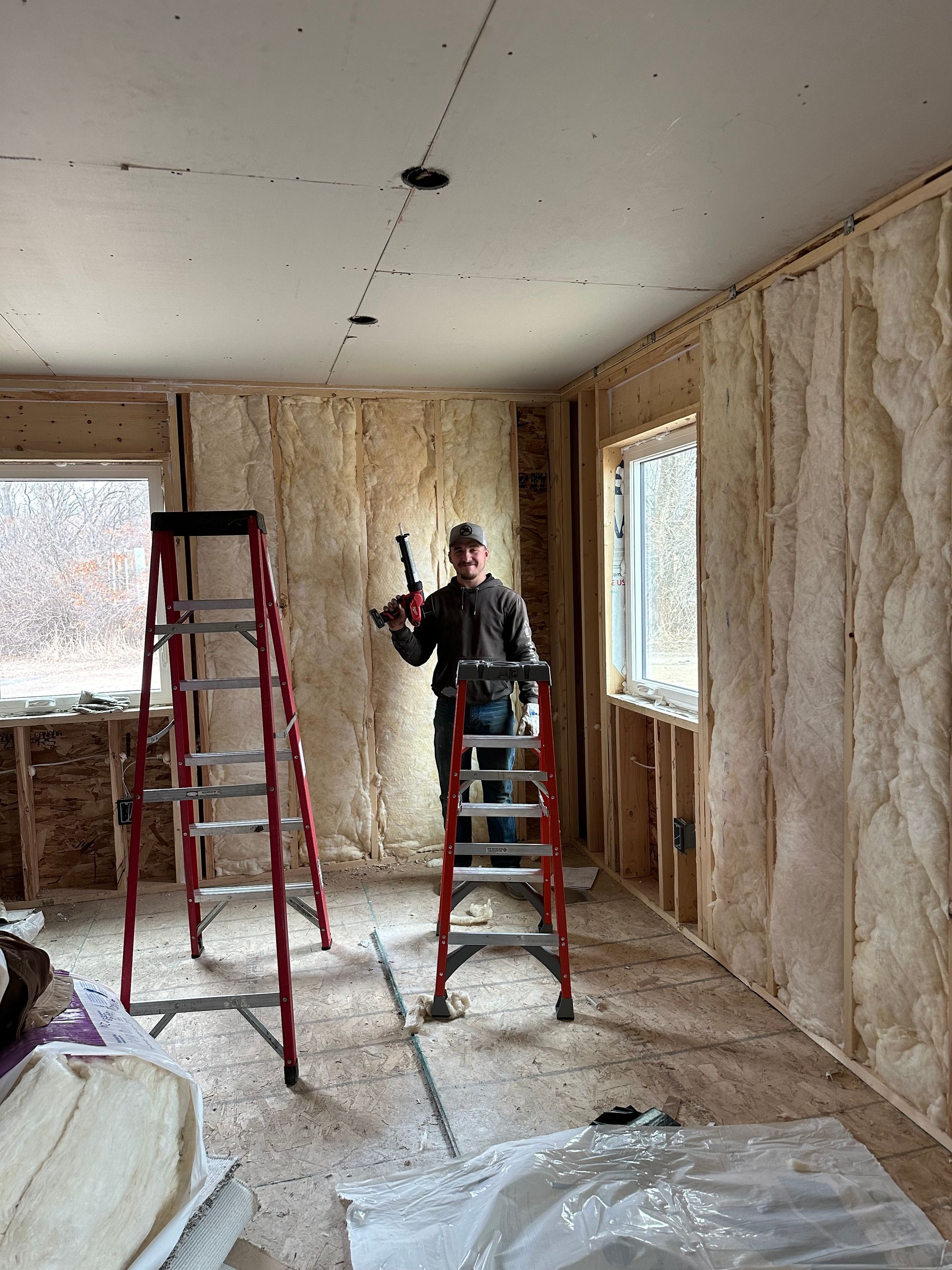 Worker standing on a ladder in an unfinished room with insulation exposed and windows on two walls.