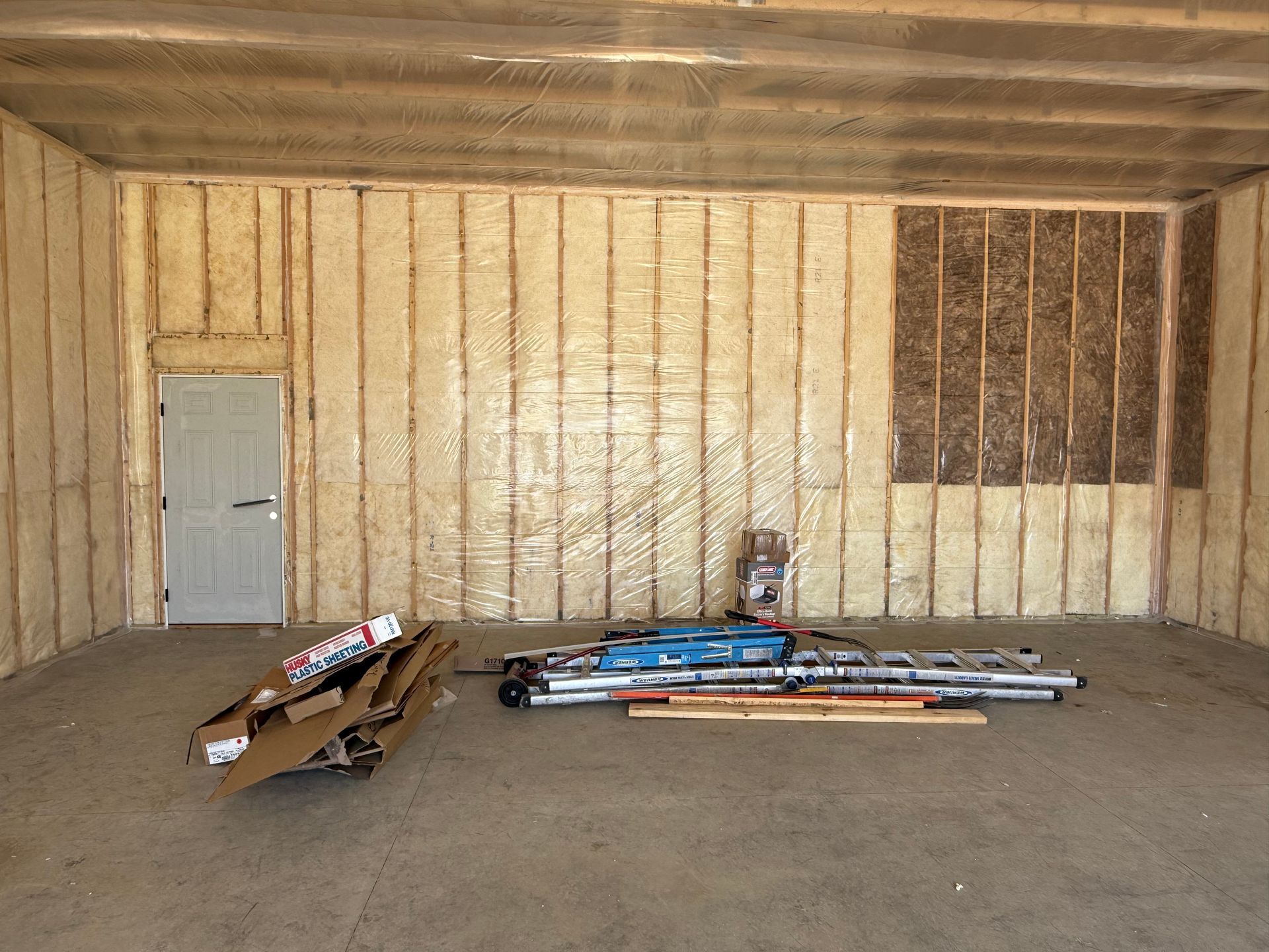Empty insulated room with stacked lumber and pipes on the floor near a gray door