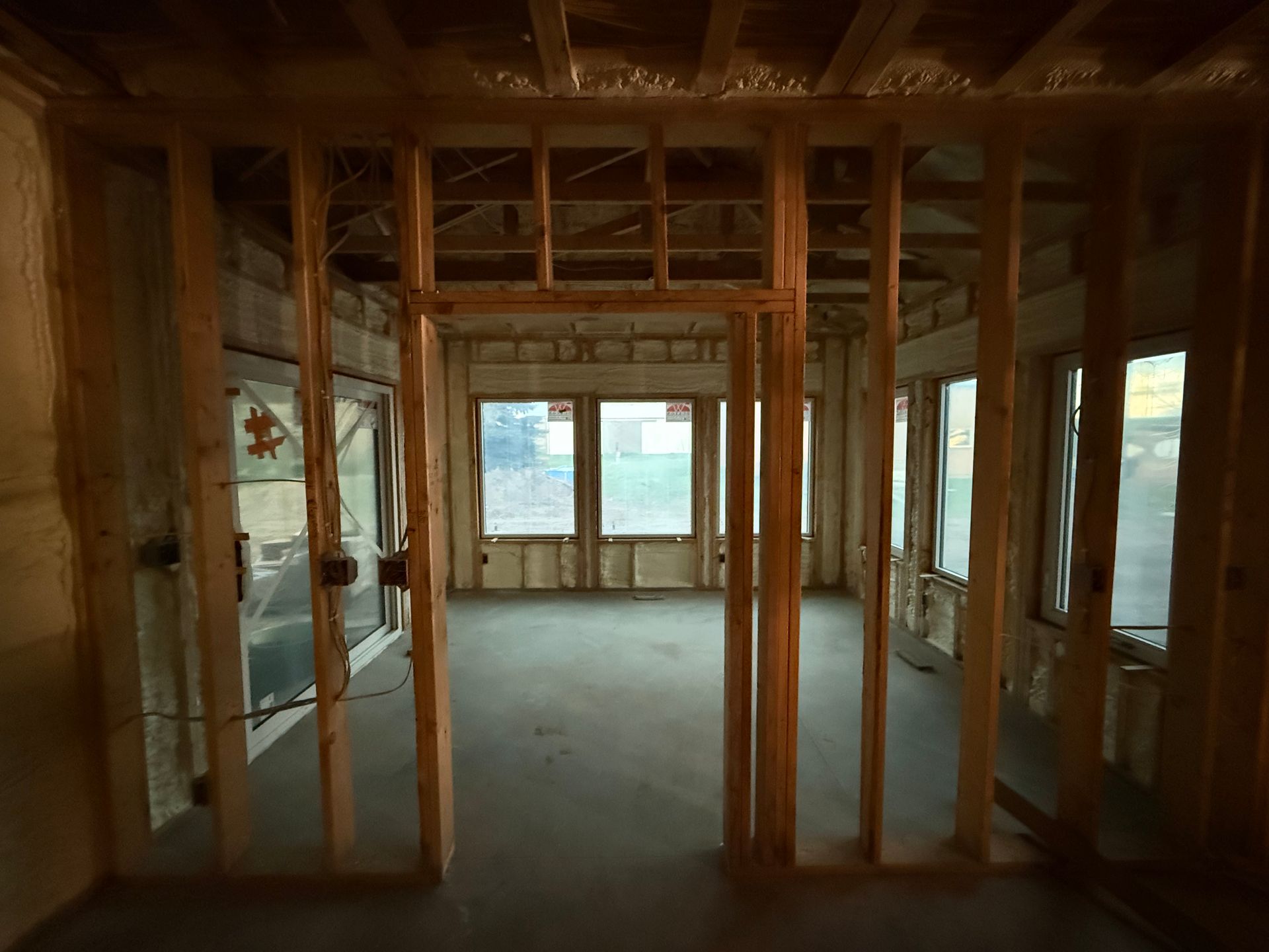 Exposed wooden framing of an unfinished room with windows and bare walls overlooking a grassy view