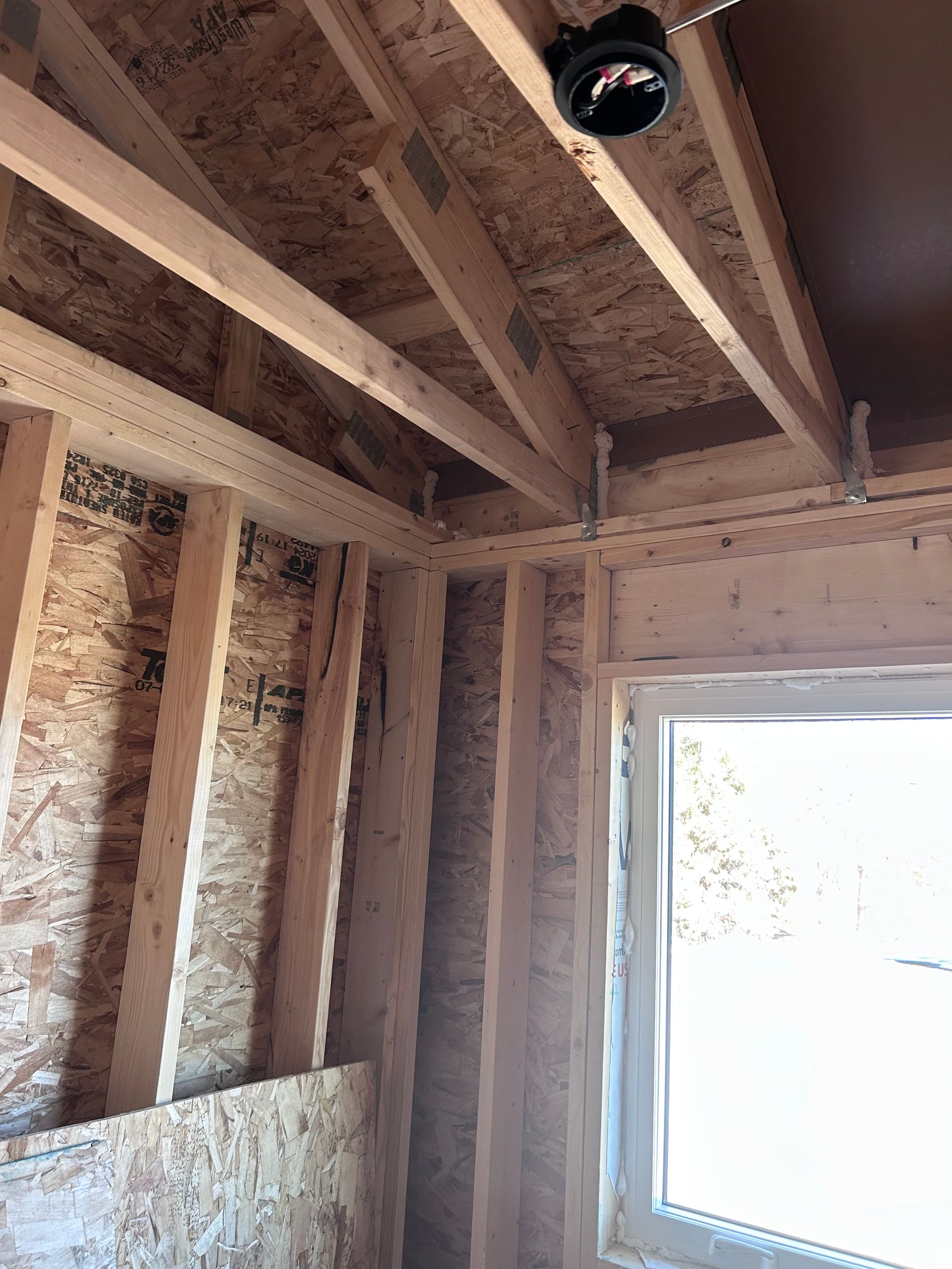 Unfinished interior wall framing with exposed insulation, roof trusses, and a window letting in bright light