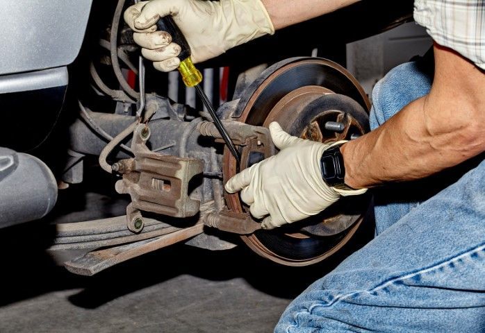 A mechanic wearing gloves uses a screwdriver while working on a vehicle's brake disc assembly in a garage.