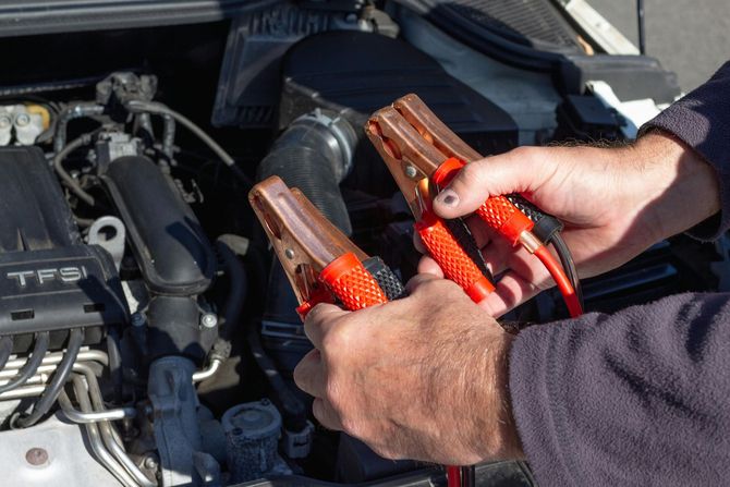 A person holding two red-handled jumper cable clamps in front of a car engine.