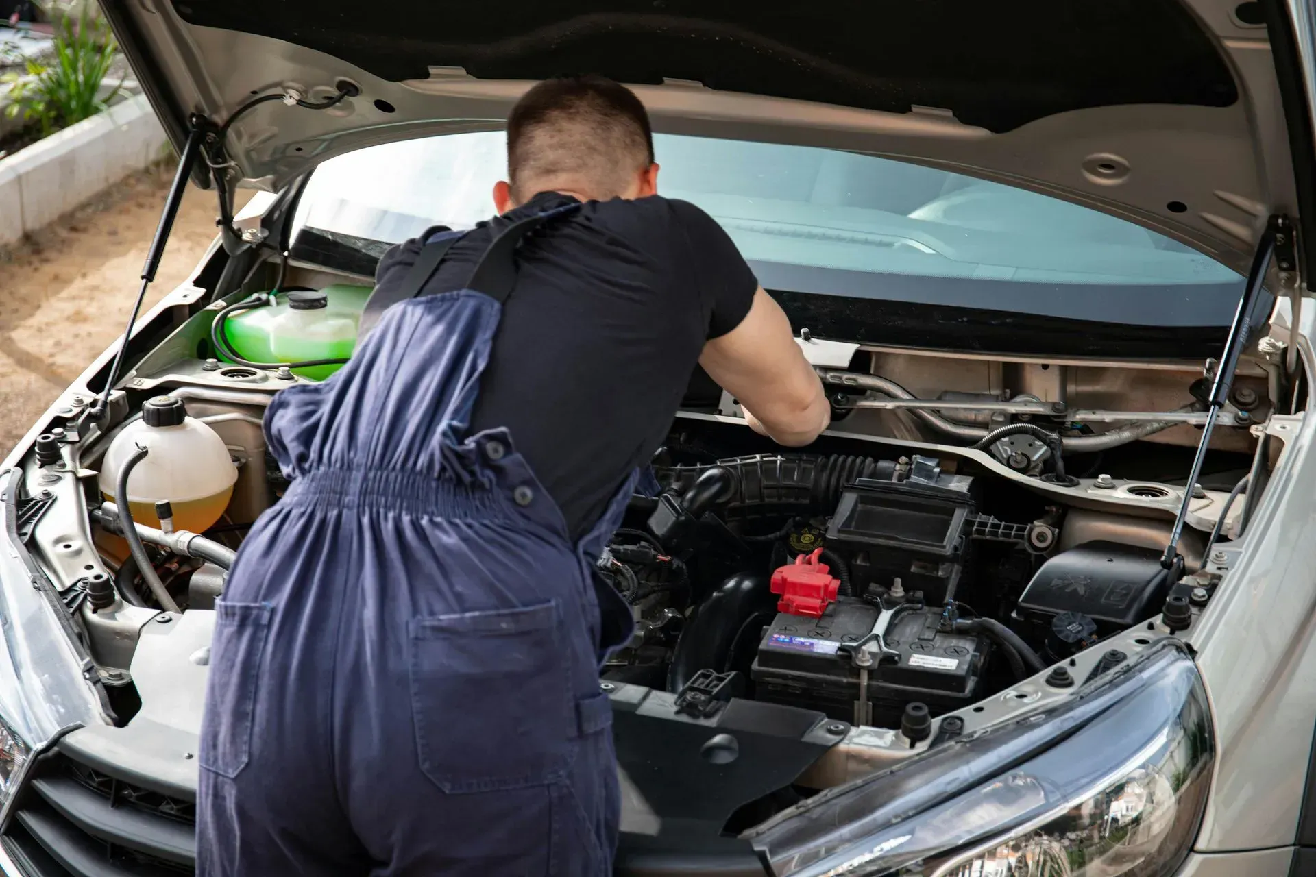 Mechanic in blue overalls repairing a car engine under the hood.