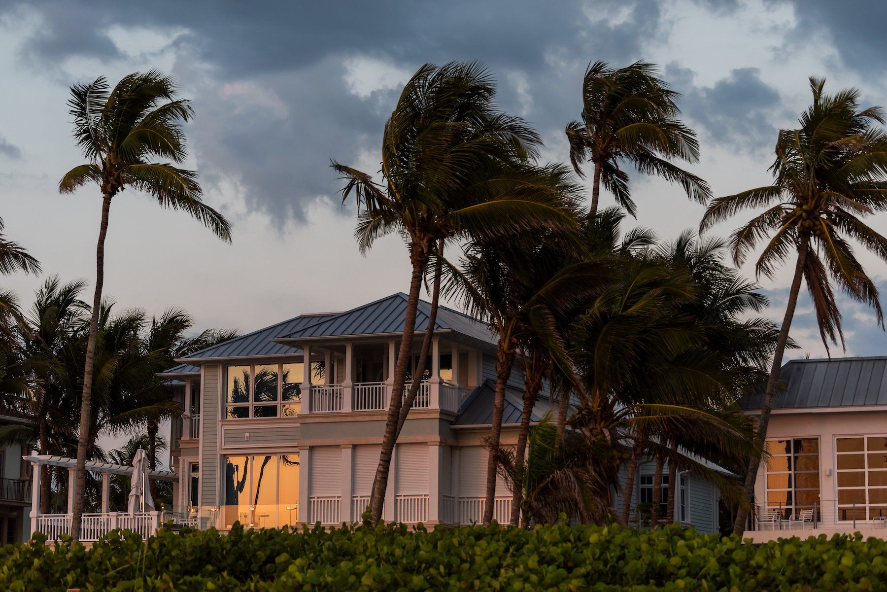 Coastal vacation home overlooking the beachfront at sunset, with stormy weather creating dramatic skies and palm trees swaying in the wind.