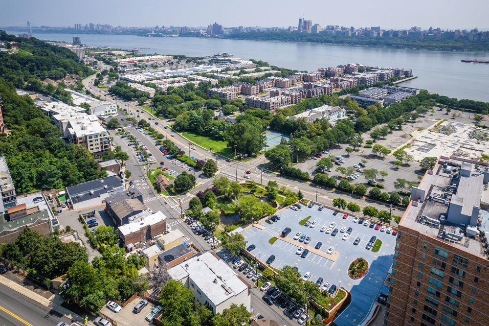 Aerial view of a town along a river. Buildings, roads, parking lots, and green spaces are visible. The cityscape is under a blue sky.