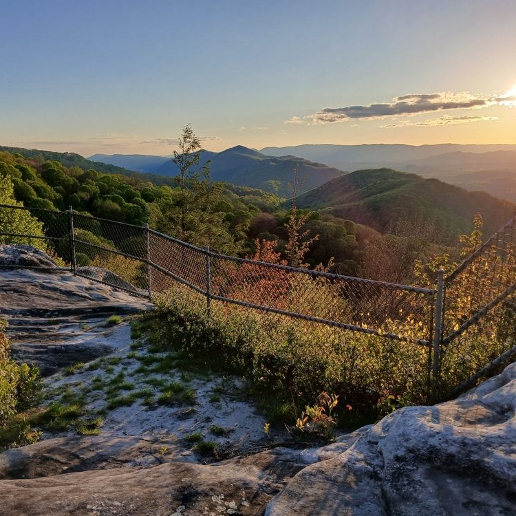 A sunset over a mountain range, viewed from a rocky, fenced-in lookout point with green and orange foliage.