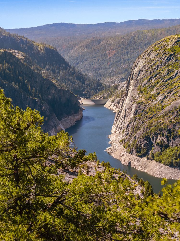 River flowing through a deep canyon, dam visible in the distance. Lush trees and rocky cliffs under a blue sky.