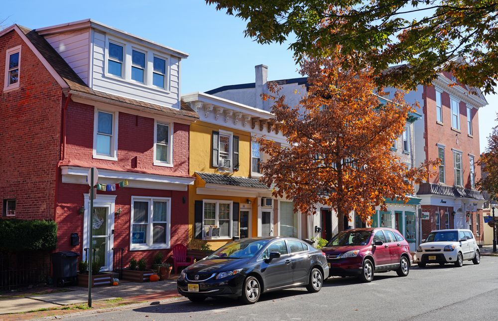 Row of colorful houses with parked cars on a street; a tree with orange leaves.