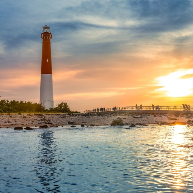 Barnegat Lighthouse stands tall by the shore during a vibrant, golden sunset, with silhouettes of people on a walkway.