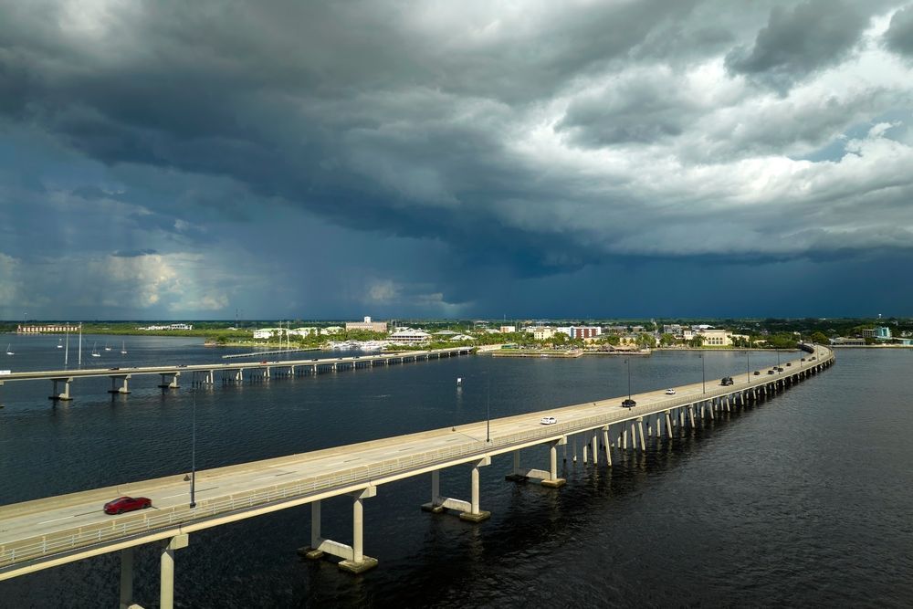 Bridge over dark water, under stormy sky with buildings on shore.
