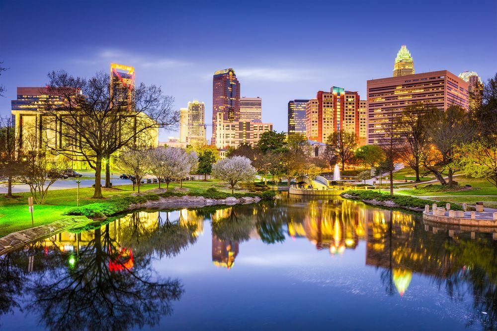Charlotte, North Carolina skyline reflected in a calm lake at dusk; buildings and trees are lit with golden light.