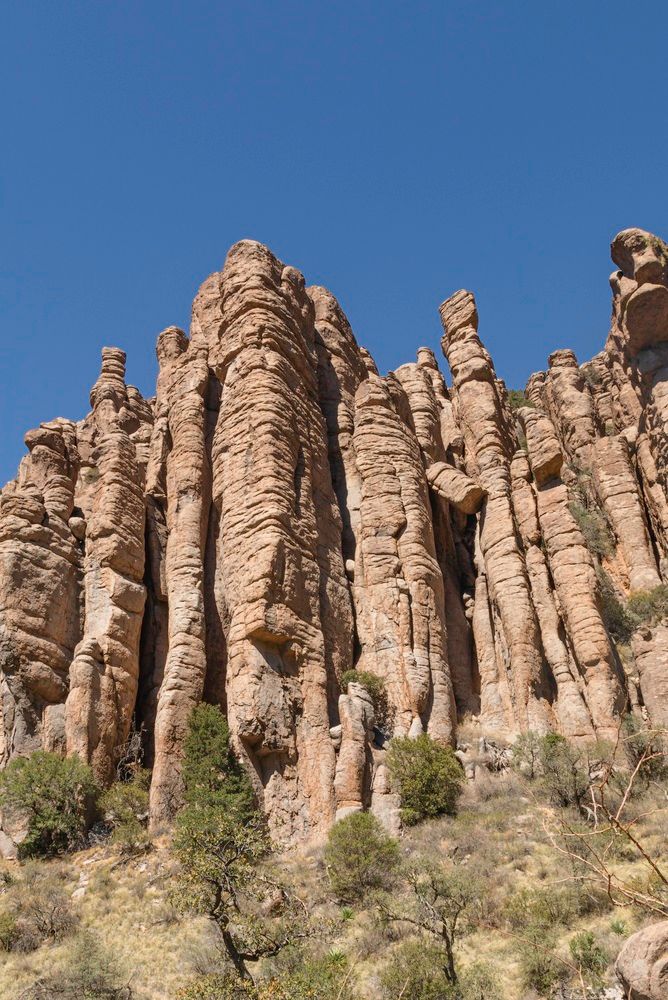 Tall tan rock spires rise against a clear blue sky with sparse shrubs below.