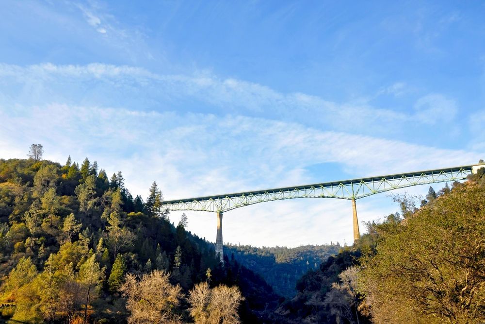 Bridge spanning a canyon, framed by trees under a blue sky with light clouds.
