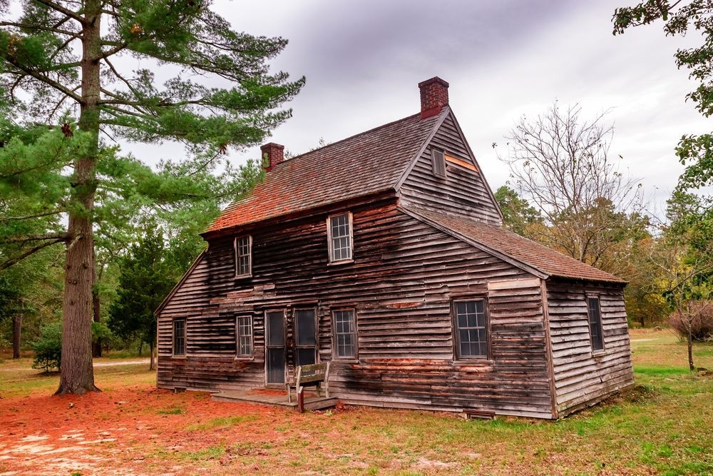 Weathered wooden house with dark brown walls, red roof, surrounded by trees. Cloudy sky.