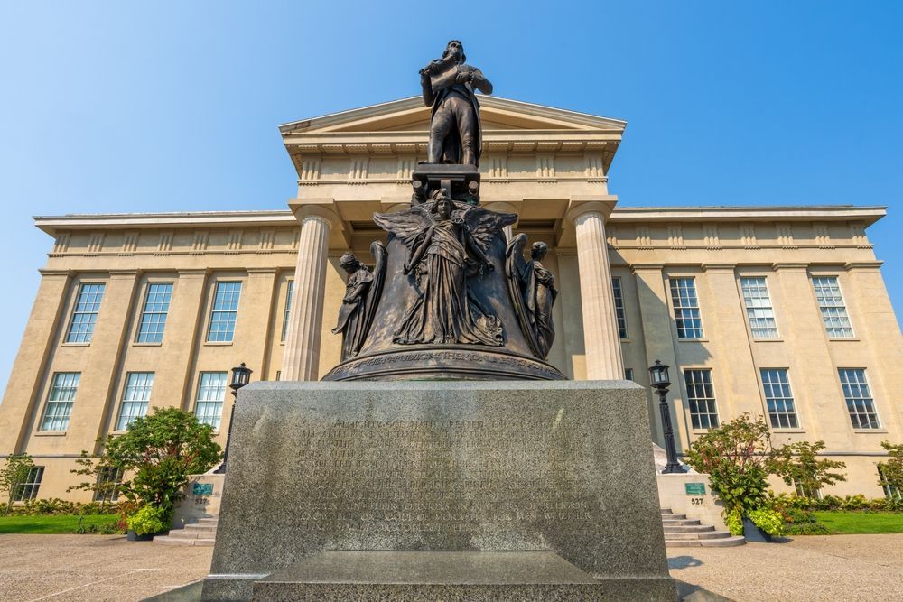 Bronze statue in front of a tan building with columns. Figures surround the statue atop a stone base, under a clear, blue sky.