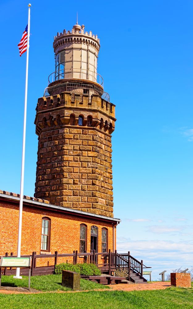 Stone lighthouse with a tower, an attached brick building, and an American flag against a blue sky.