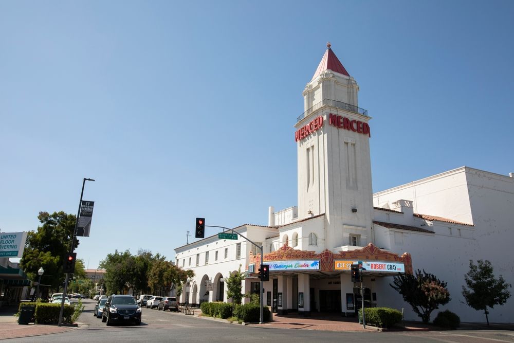 Merced Theatre, a white Art Deco building with a tall tower and red accents, on a sunny street with cars and trees.
