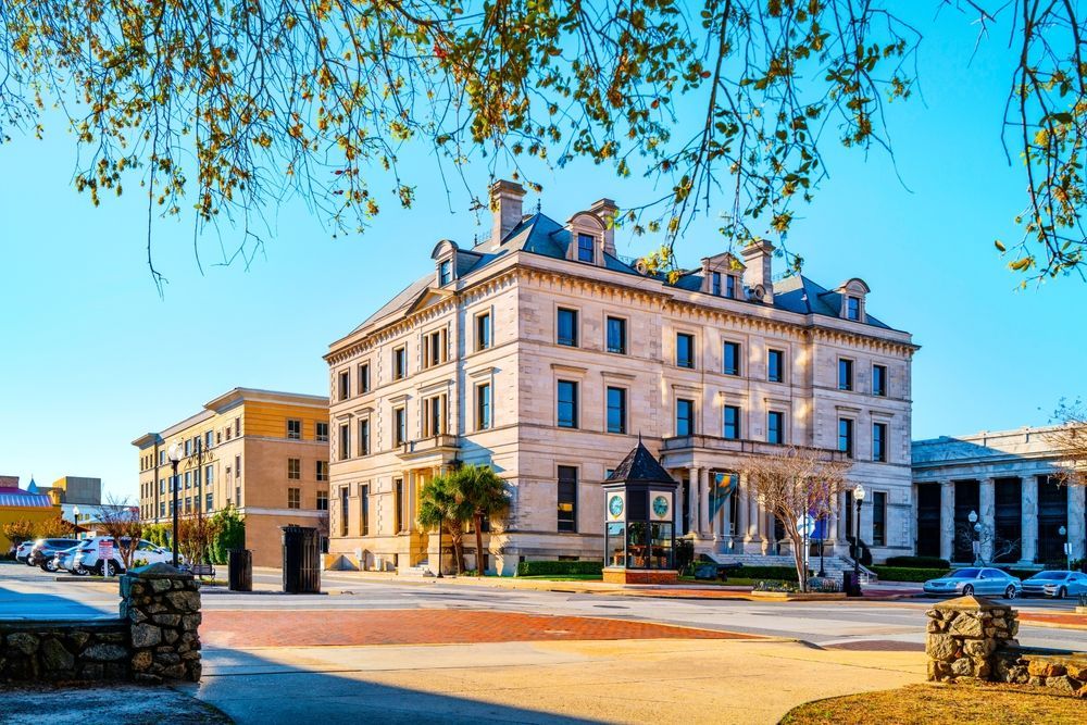 Large stone building in a city square under a blue sky, surrounded by other buildings and trees.
