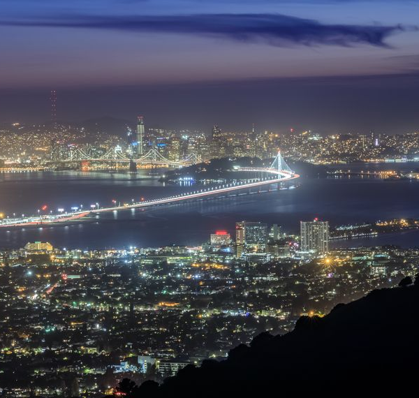 Night view of San Francisco Bay area, illuminated city lights, Golden Gate Bridge, and dark silhouette of hills.