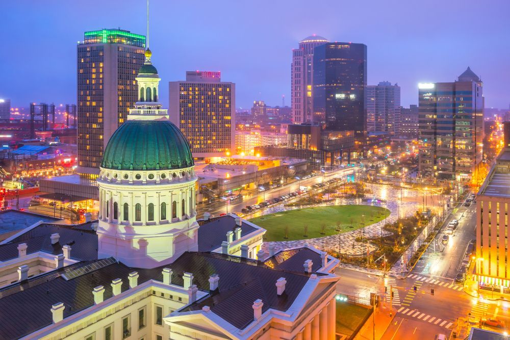 City skyline at dusk with the dome of a historic building in foreground. Lights illuminate the buildings and streets.
