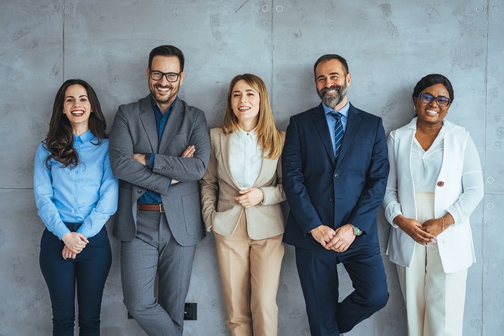 Five diverse businesspeople in formal attire lean against a grey wall, smiling.