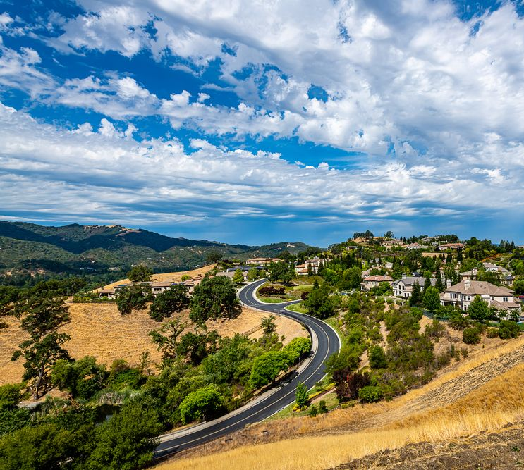 Rolling hills with a winding road leading to a residential area under a cloudy blue sky.