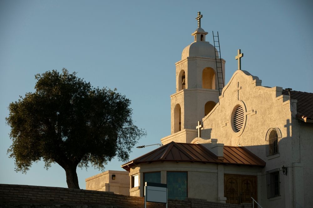 Church building with a bell tower and cross on a sunny day; a tree stands in front.