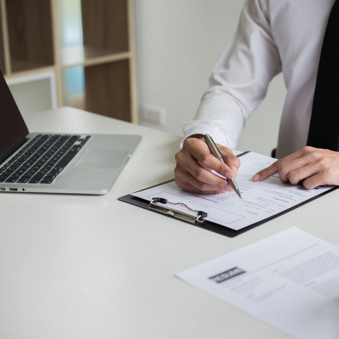 Person in a white shirt writing on a document at a desk, laptop to the left.