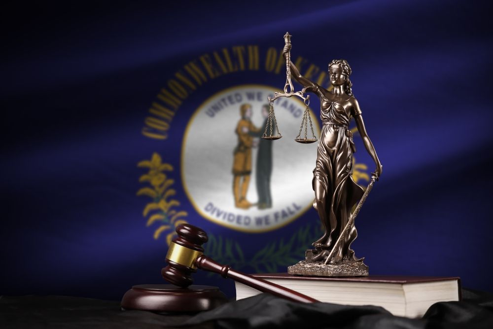 A statue of lady justice sits on top of a book in front of a commonwealth of kentucky flag