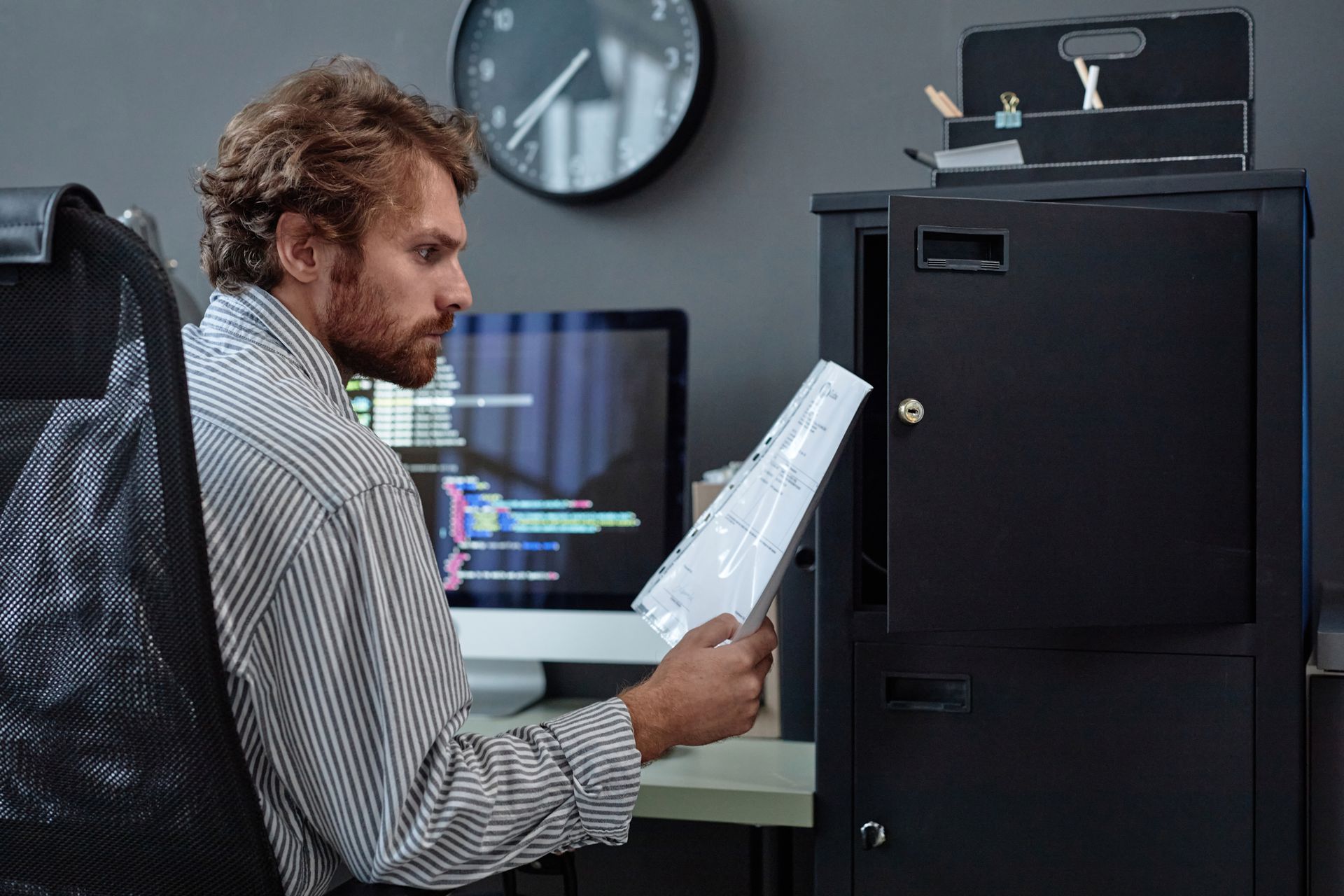 A man is looking at a piece of paper in front of a computer sitting next to a locking file cabinet for sensitive files regarding cybersecurity for CRAs