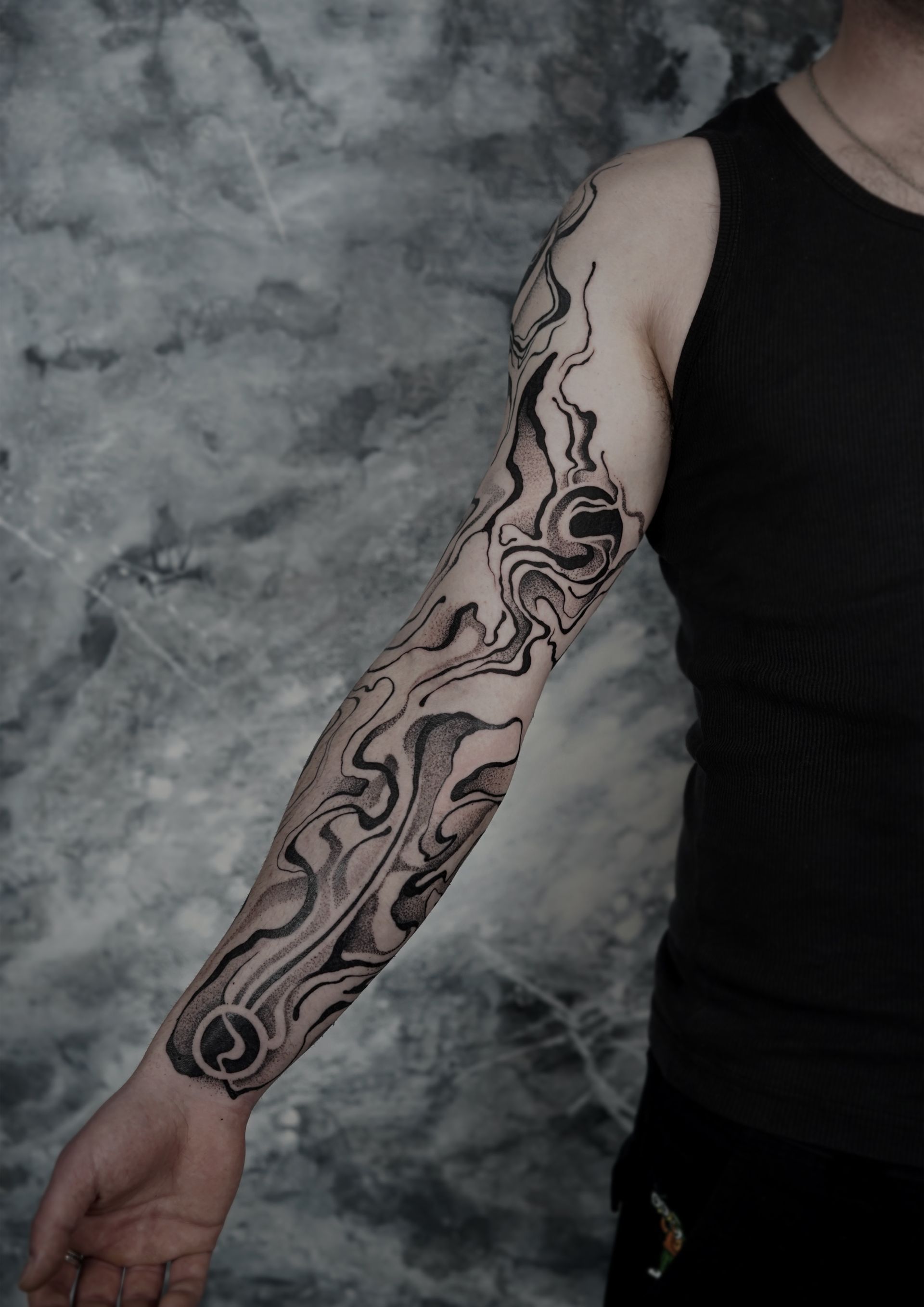 Person with full-sleeve tattoo on left arm; swirling, abstract black ink design; wearing black tank top.