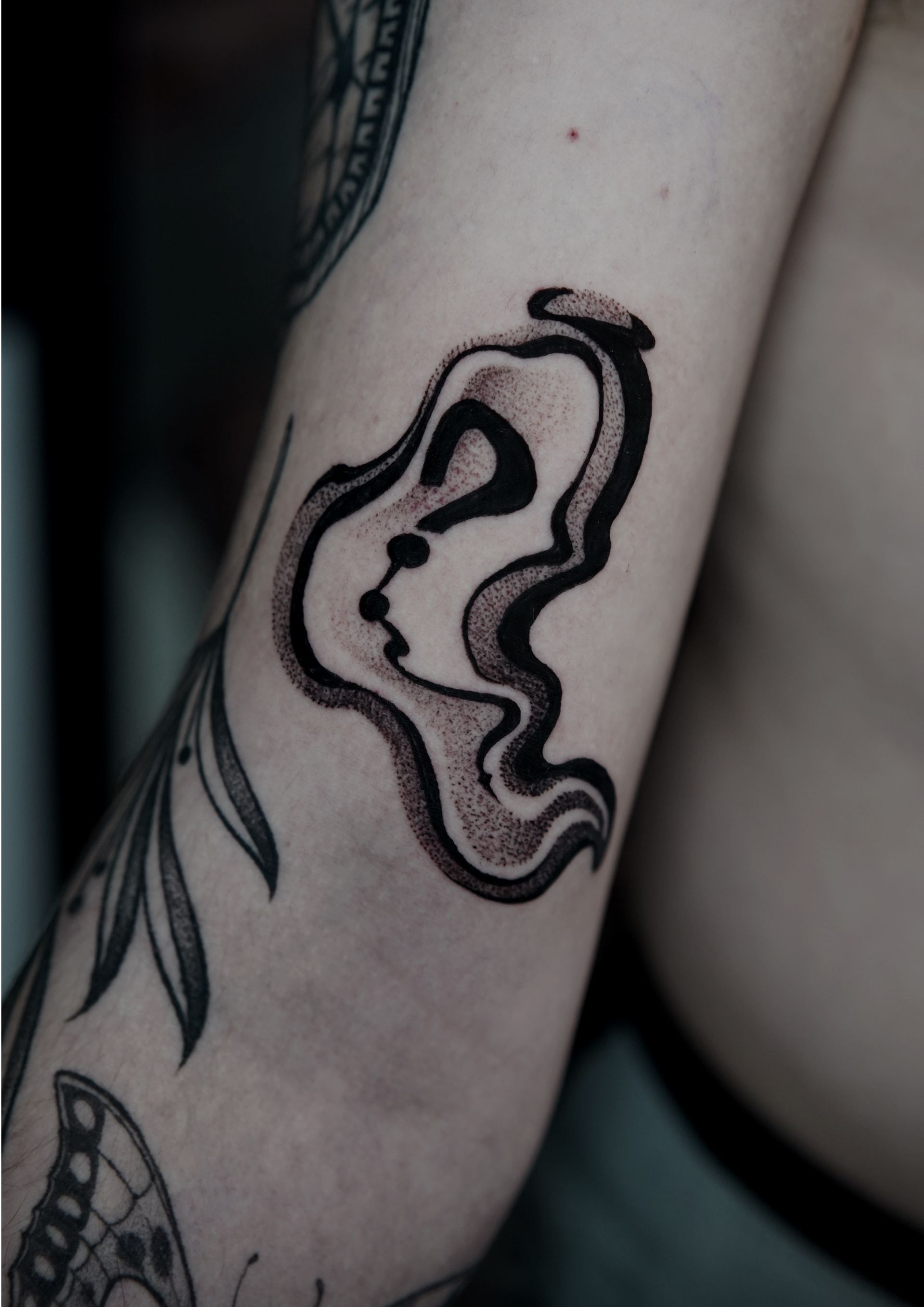 Black tattoo on arm; abstract melting design with a profile and question mark shape.