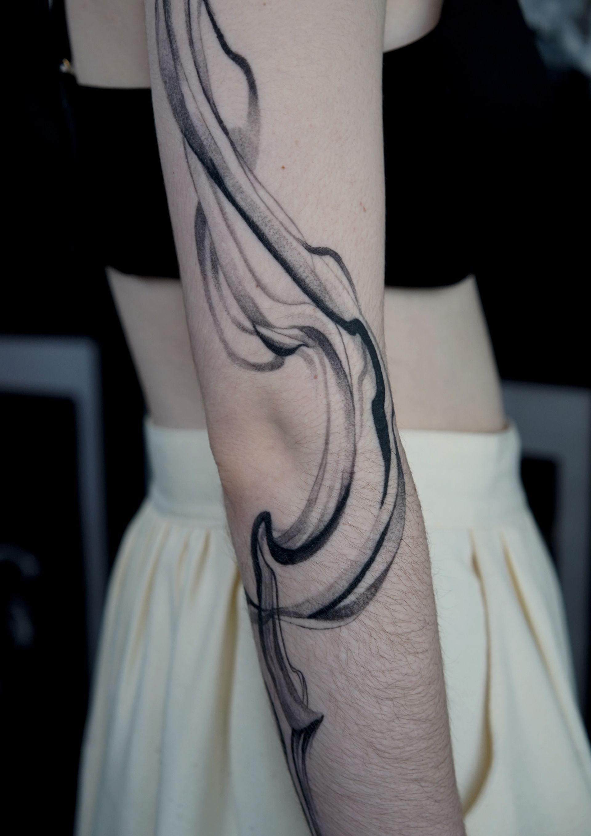 Arm with flowing, abstract black ink tattoo, person in black top and cream skirt.