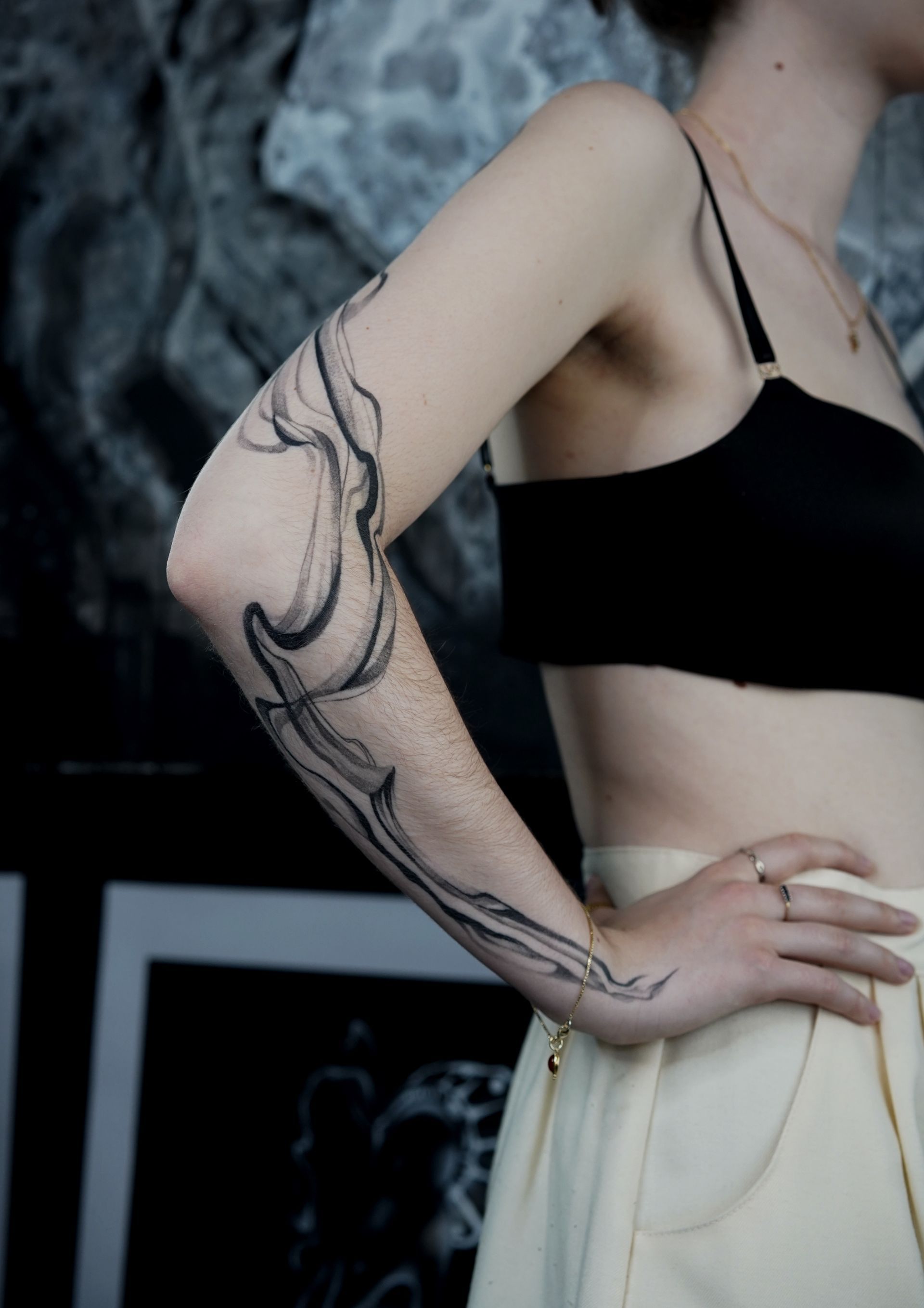 Woman with black arm tattoo, wearing a black top and tan pants, hand on hip.