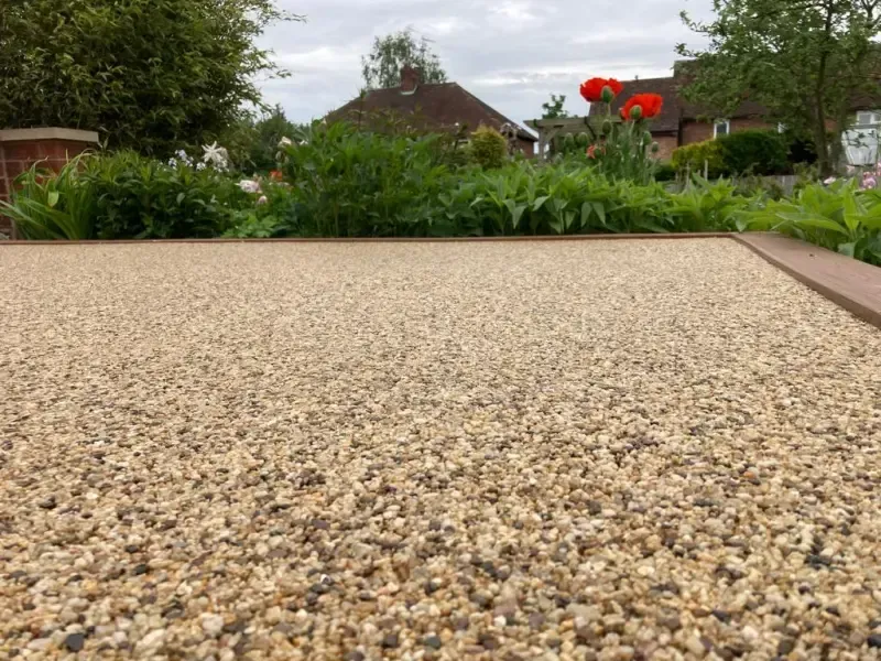 Close-up view of light brown pebble-covered pathway leading to a garden with red flowers and green foliage.