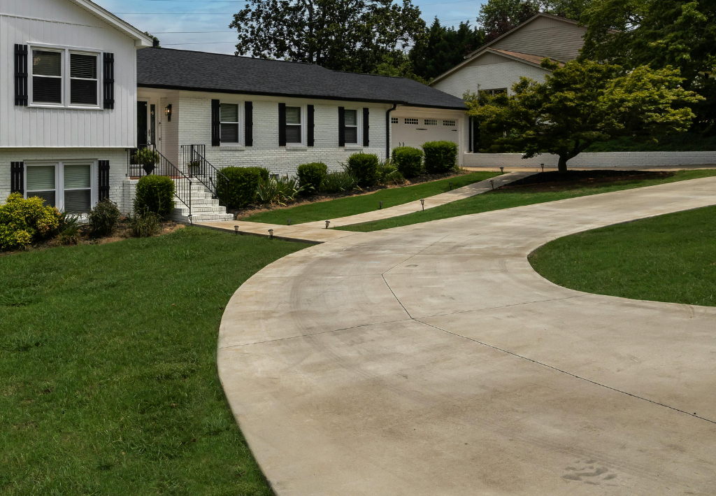 White house with black shutters, driveway curves, green lawn, trees, and blue sky.