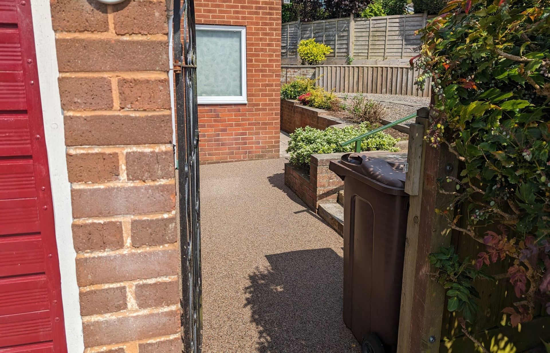 View of a gravel pathway leading to a garden. Brown trash bin, brick wall, and red-painted structure visible on the left.