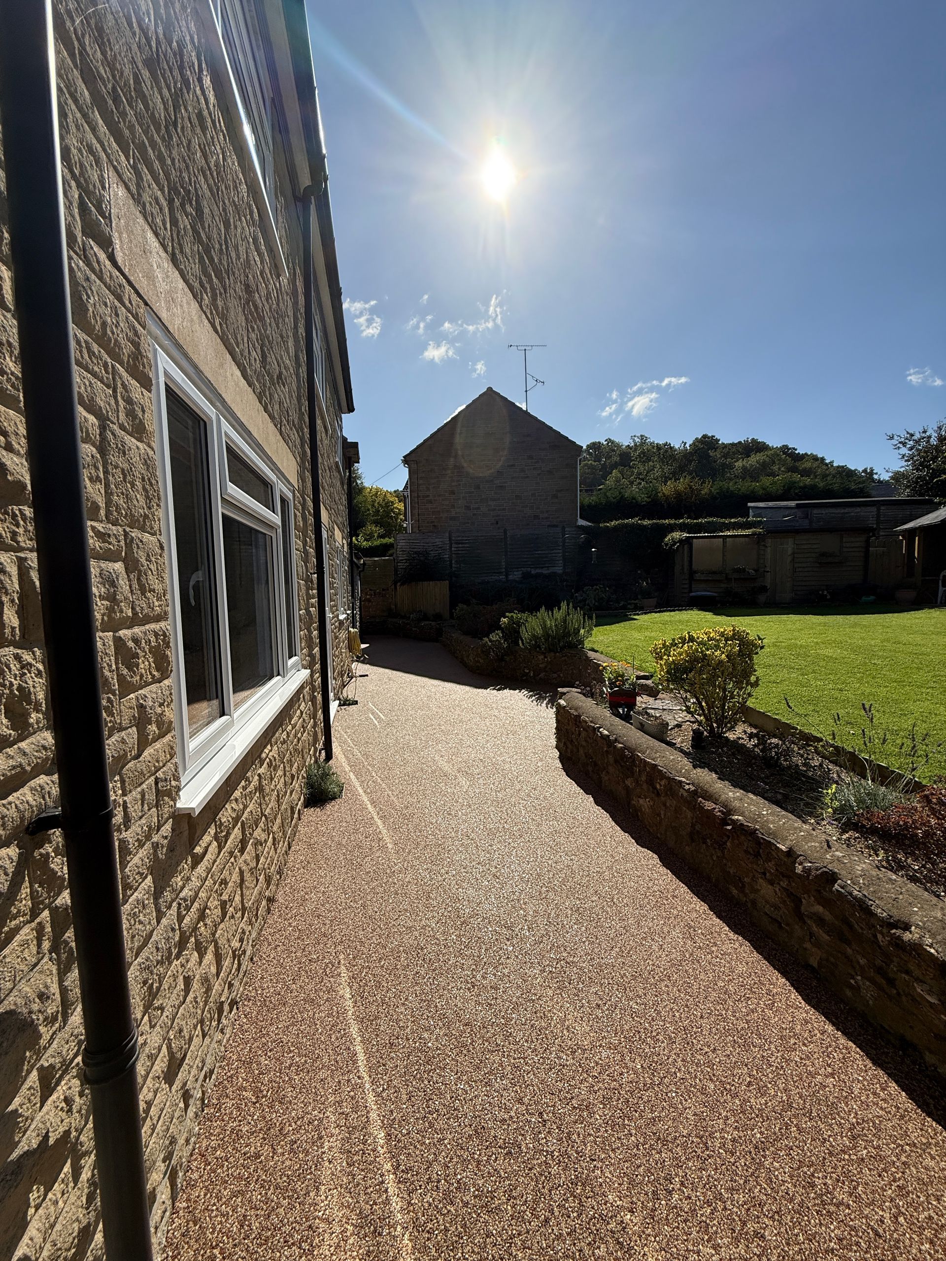 A stone path next to a stone building under a bright sun. Green grass and a small building are further in the background.