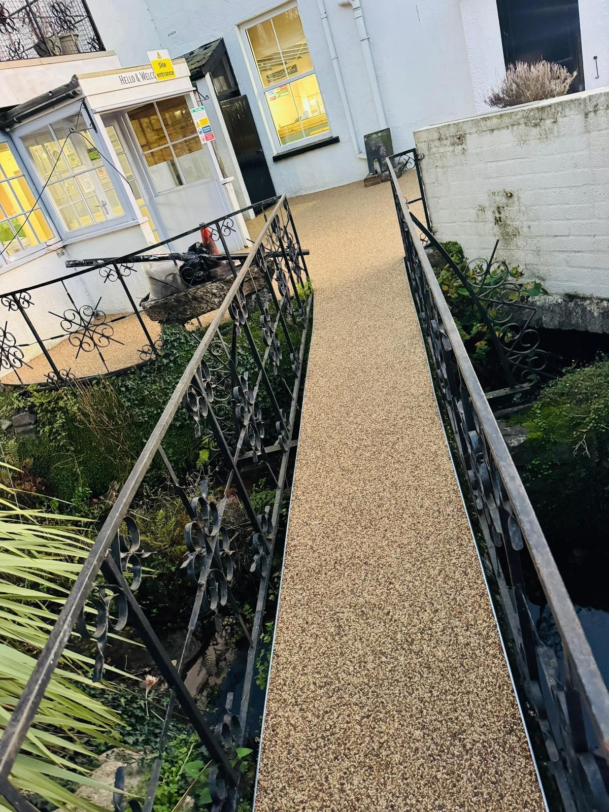 Narrow pedestrian bridge with decorative railings over a water feature, leading to a white building.