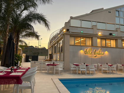 Terrazza esterna di un caffè al tramonto con tavolini rossi e bianchi, palme e un'insegna luminosa dell'edificio che si riflette in una piscina.