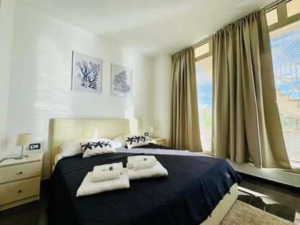 Luminosa camera da letto con letto, tende beige e finestre a tutta altezza con vista sull'esterno.