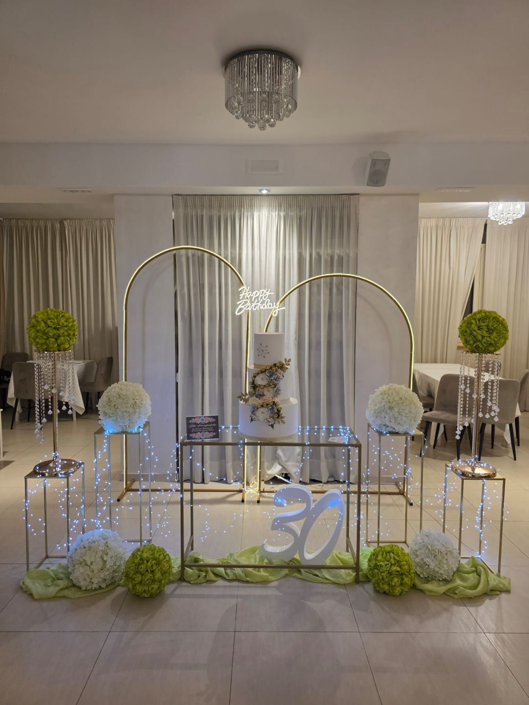 Elegante allestimento per la festa del 30° compleanno in bianco e oro con archi di palloncini, fiori e un tavolo di vetro.