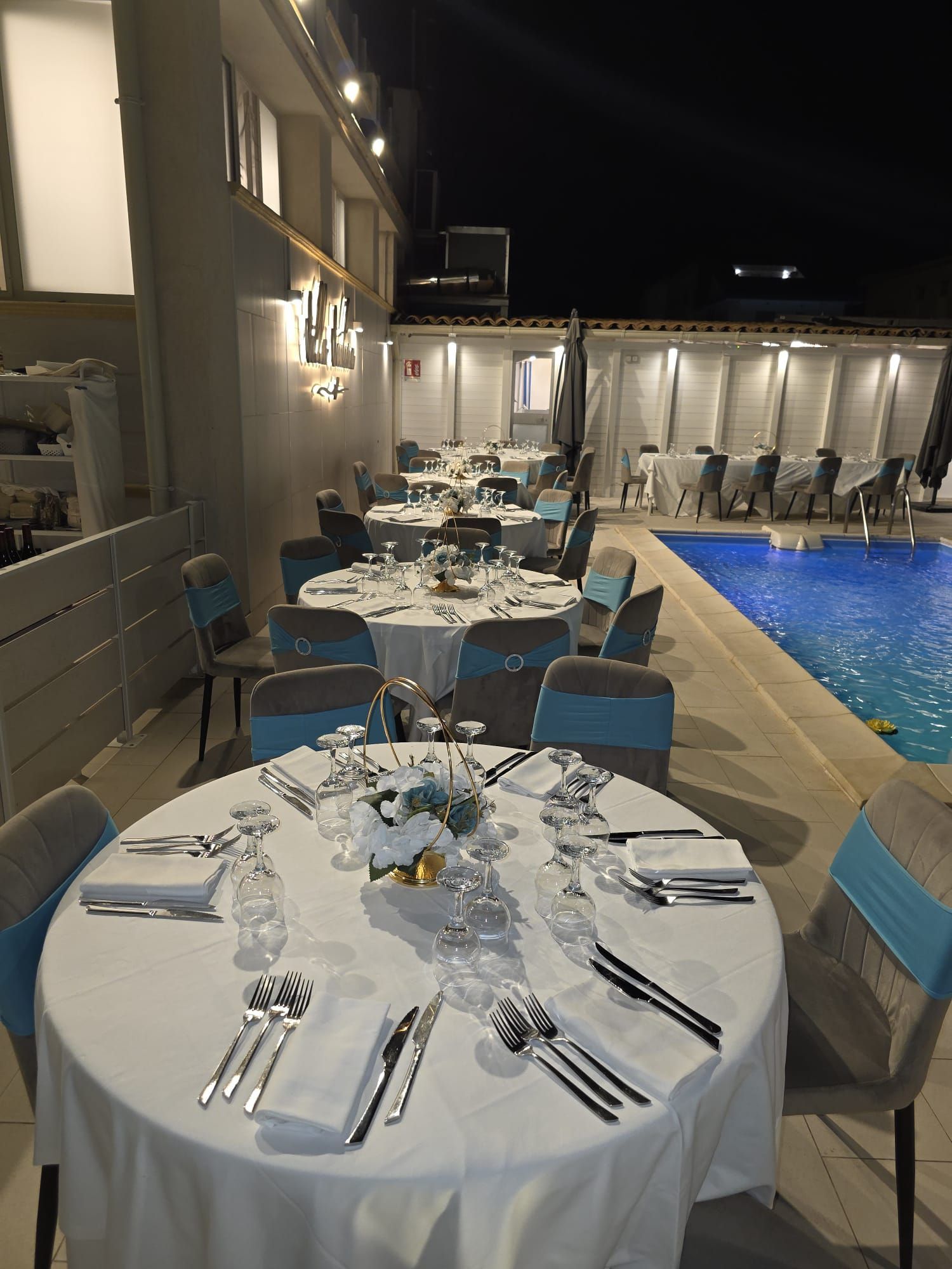 Tavoli da pranzo all'aperto accanto a una piscina illuminata di notte, apparecchiati con tovaglie bianche e sedie blu.